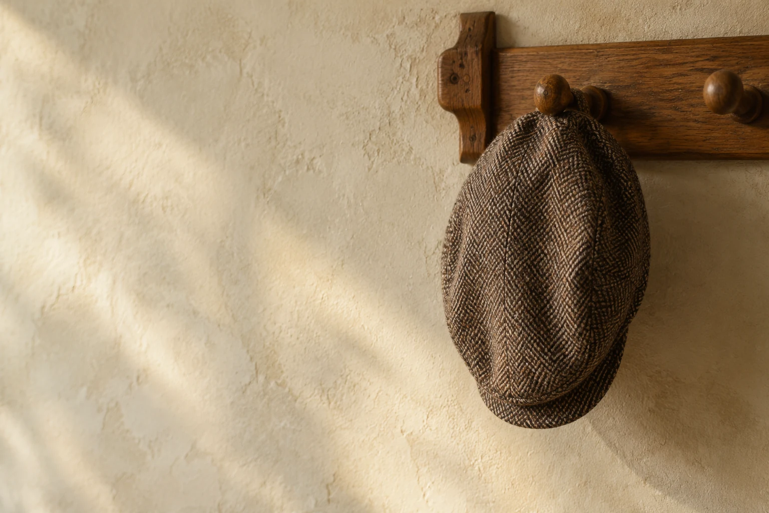 Brown herringbone tweed flat cap hanging on a wooden peg against a pale plaster wall